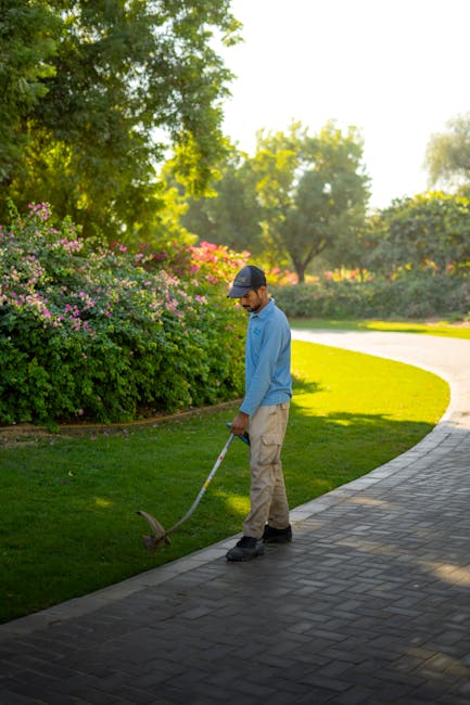 A garden maintenance worker wearing a blue shirt, beige trousers, and a dark baseball cap is using a strimmer to trim the edge of a well-maintained lawn along a paved pathway. The garden features lush