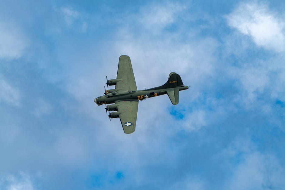 A vintage military aircraft, specifically a World War II-era bomber, flying low against a partly cloudy blue sky. The aircraft is painted in a light gray camouflage with distinctive markings, includin