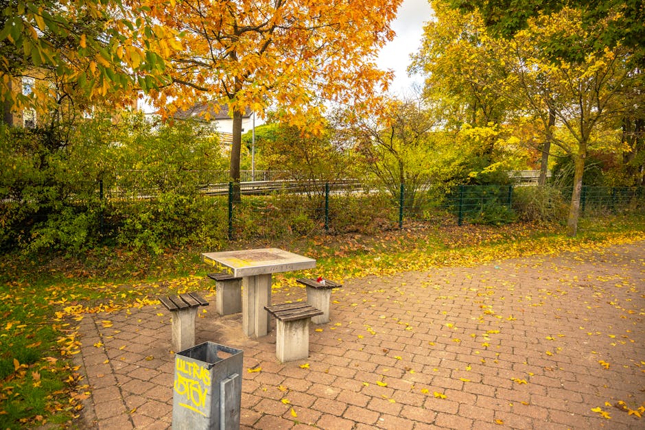 A small outdoor area featuring a concrete picnic table with three matching concrete stools positioned around it, situated on a brick-paved surface scattered with fallen yellow and brown autumn leaves.