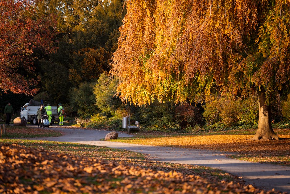 A small outdoor area featuring a concrete picnic table with three matching concrete stools positioned around it, situated on a brick-paved surface scattered with fallen yellow and brown autumn leaves.